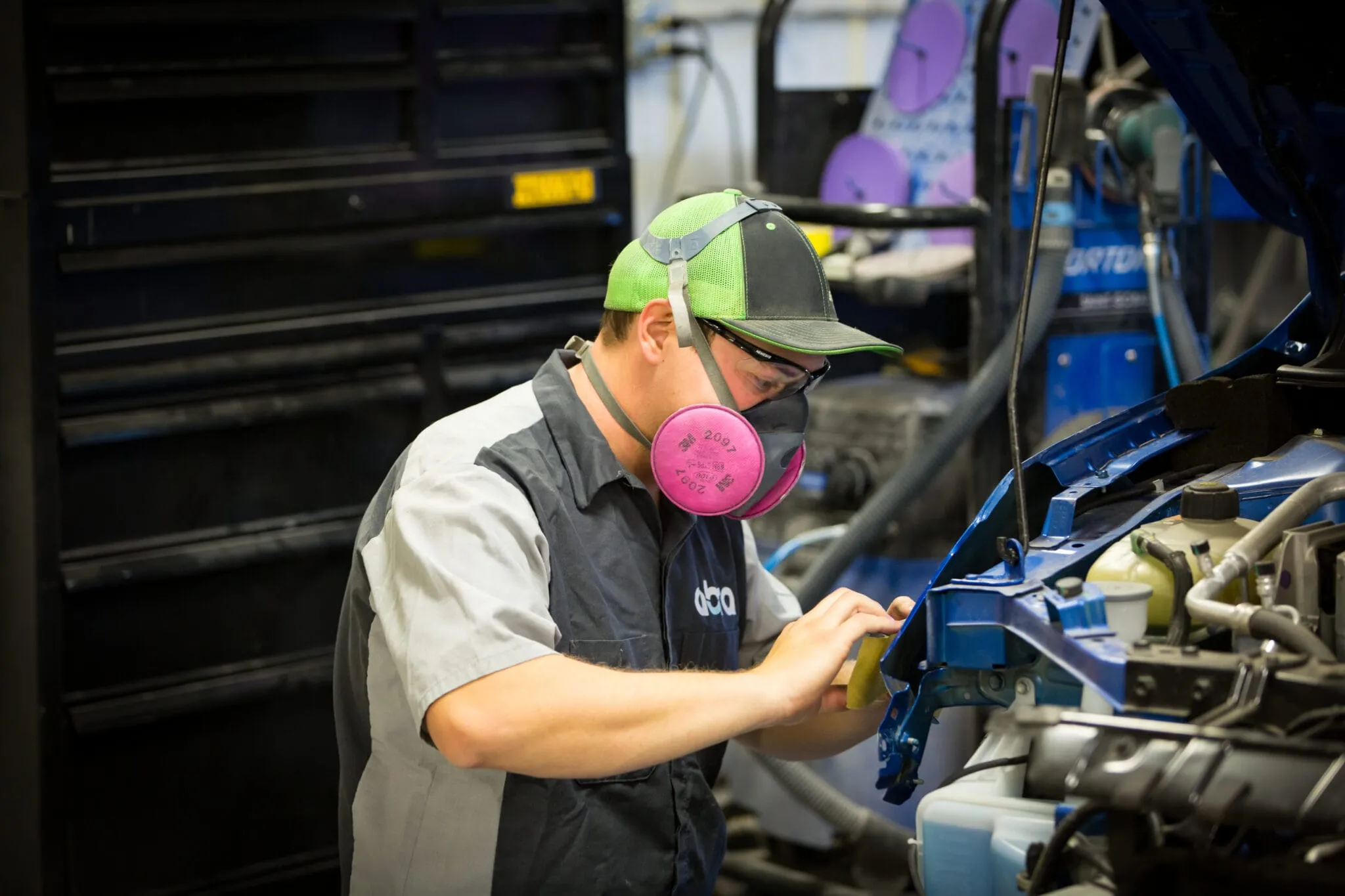 Technician in a cap and respirator working on a blue vehicle with the hood open