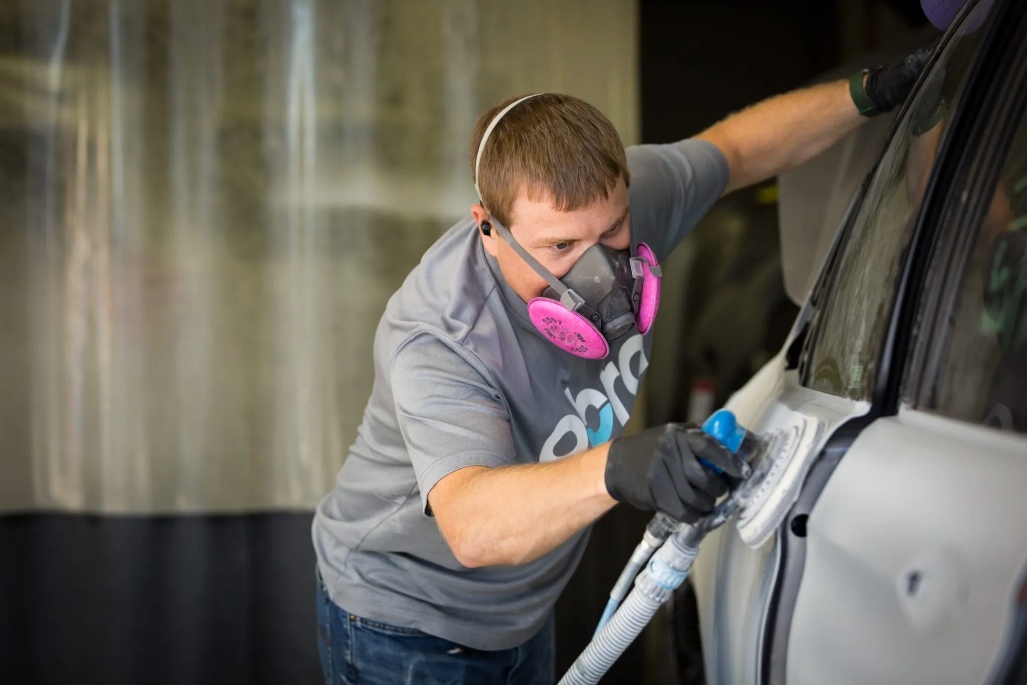 Technician in a respirator and ABRA shirt sanding a vehicle door panel